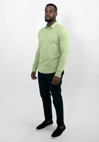 Lichtgroene lange mouwen polo met een kraag, gecombineerd met donkerblauwe broek en zwarte instapschoenen, staand tegen een witte achtergrond.