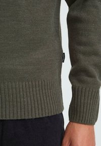 Close-up of olive green ribbed knit sweater sleeve and hem with black woven brand tag, worn with dark pants.