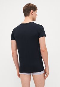 Joven de espaldas vistiendo una camisa azul marino de manga corta y pantalones cortos gris claro sobre un fondo blanco liso.