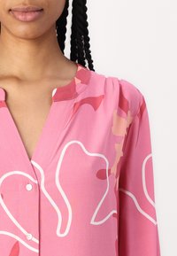Rosa Bluse aus gewebtem Stoff mit V-Ausschnitt, weißen abstrakten Mustern und Knopfdetails auf der Vorderseite. Glatte Textur.