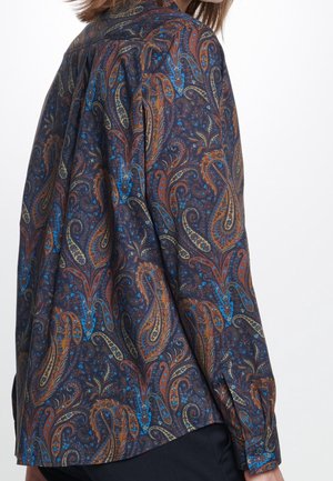 Personne portant une chemise à manches longues avec un motif paisley sombre dans des tons bleus, marron et orange, vue de dos et de côté.