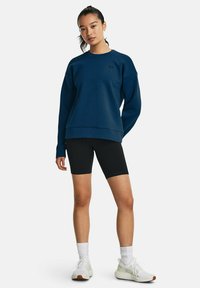 Felpa blu navy con vestibilità rilassata e collo rotondo, abbinata a pantaloni shorts da biker neri. Il modello indossa anche scarpe e calze sportive bianche.