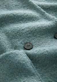 Světle modrá texturovaná látka s měkkým, chlupatým vzhledem, zdobená tmavě šedými knoflíky se dvěma otvory, uspořádaná na zmuchlaném povrchu.