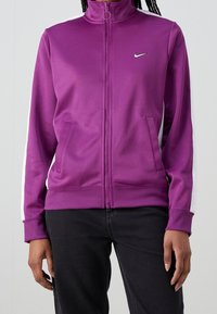 Lila Nike jacka med dragkedja, hög krage, sidosfickor och vita detaljstreck på ärmarna. Slät tyg med figursydd passform.