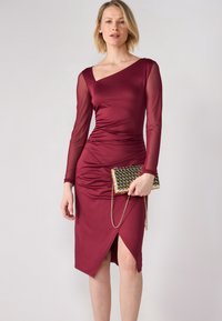 Burgunderfarget tettsittende kjole med lange gjennomsiktige ermer og asymmetrisk halslinje. Har samlet tekstur og en sideslitt, parret med en gull clutch.