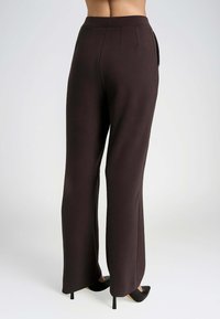 Pantaloni marroni a gamba larga con una texture liscia, dotati di una vita alta, pieghe frontali e tasche laterali. Abbinati a tacchi a punta neri.