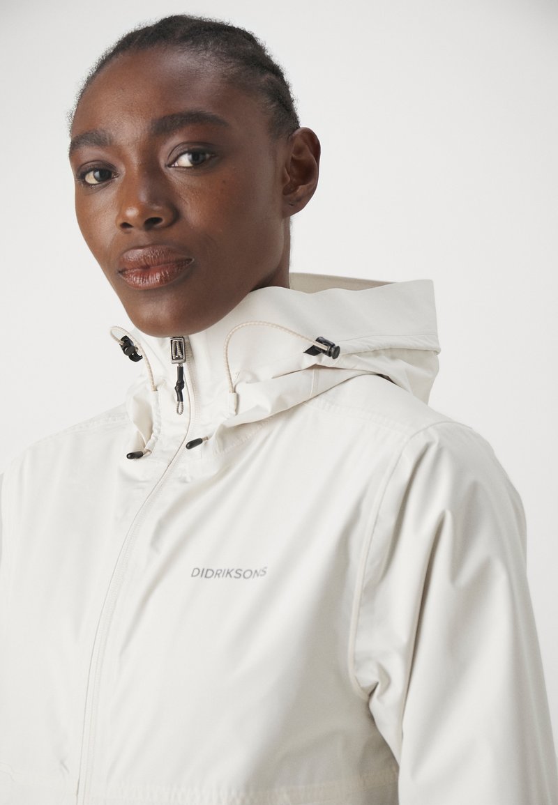 Cremfarbene, wasserdichte Jacke mit hohem Kragen, verstellbarem Kapuzenverschluss und Reißverschluss auf der Vorderseite; mit dem Markenlogo auf der Brust.