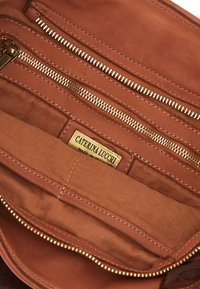 Interno della borsa in pelle tan con molteplici scomparti con zip. Presenta hardware dorato, fodera testurizzata e un'etichetta del marchio.