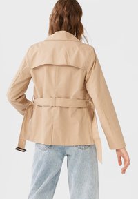 Béžový trench coat s páskem v pase, detailem na zadním díle a dlouhými rukávy. Vyroben z hladké látky, ve volném střihu.