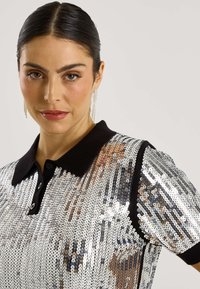 Polo de lentejuelas plateadas con cuello negro, diseño ajustado y mangas cortas, destacando una textura lisa y reflectante.