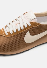 Baskets de sport marron et beige avec une tige en tissu texturé, lacets blancs et un logo swoosh beige sur le côté. Semelle extérieure en caoutchouc.