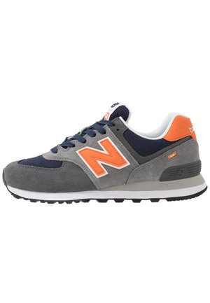Grijze en marineblauwe sneaker met oranje "N" logo, witte zool en zwarte loopzool, gezien vanaf de buitenkant.