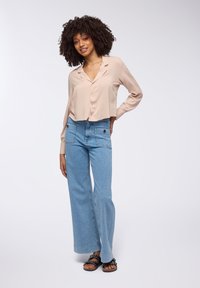 Blusa cropped beige chiaro con bottoni abbinata a jeans a gamba larga blu chiaro con due tasche frontali, indossata con sandali neri.