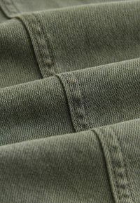 Tela verde caqui con una fina textura de sarga, con detalles de costura y capas dobladas. El material parece resistente y duradero.