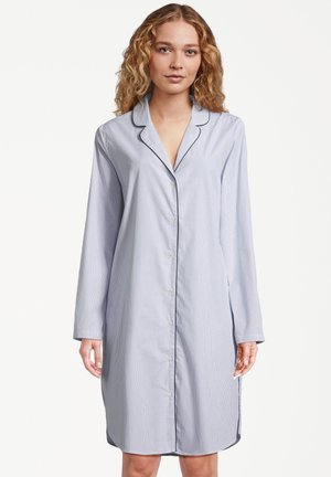 Donna con capelli biondi ricci che indossa una camicia da notte blu chiaro, a pulsanti, di lunghezza fino al ginocchio, con rifiniture scure e maniche lunghe.
