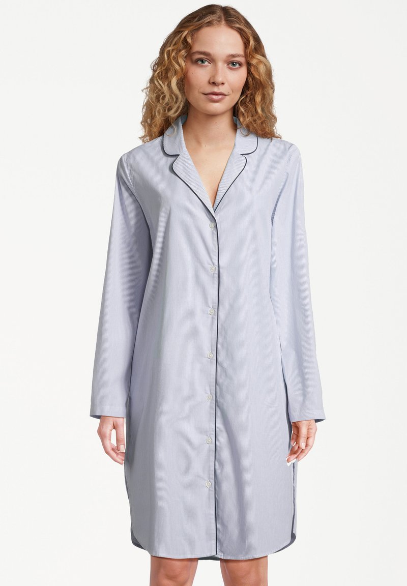 Kobieta z kręconymi blond włosami, ubrana w nocną koszulę do kolan w jasnoniebieskim kolorze z ciemnym wykończeniem i długimi rękawami.