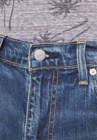 Mörkblå denimjeans med gula stygn, metallknapp och bälteloopar. Grå t-shirt med ett avslappnat palmbladsmönster överst.