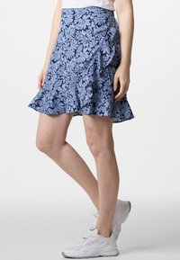 Gonna a portafoglio floreale in tessuto blu navy con fiori azzurri, orlo arricciato e tasca laterale. Indossata con scarpe da ginnastica bianche.