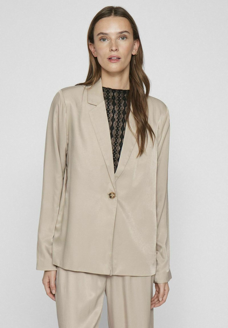 Blazer de satén beige con un cierre de un solo botón, solapas con muesca y mangas largas. Combinado con una blusa de encaje negra por debajo.