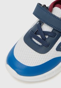 Zapatillas de malla blancas con acentos sintéticos azules y una correa de Velcro color marino. Presenta material texturizado, cordones planos y una forma de punta redondeada.