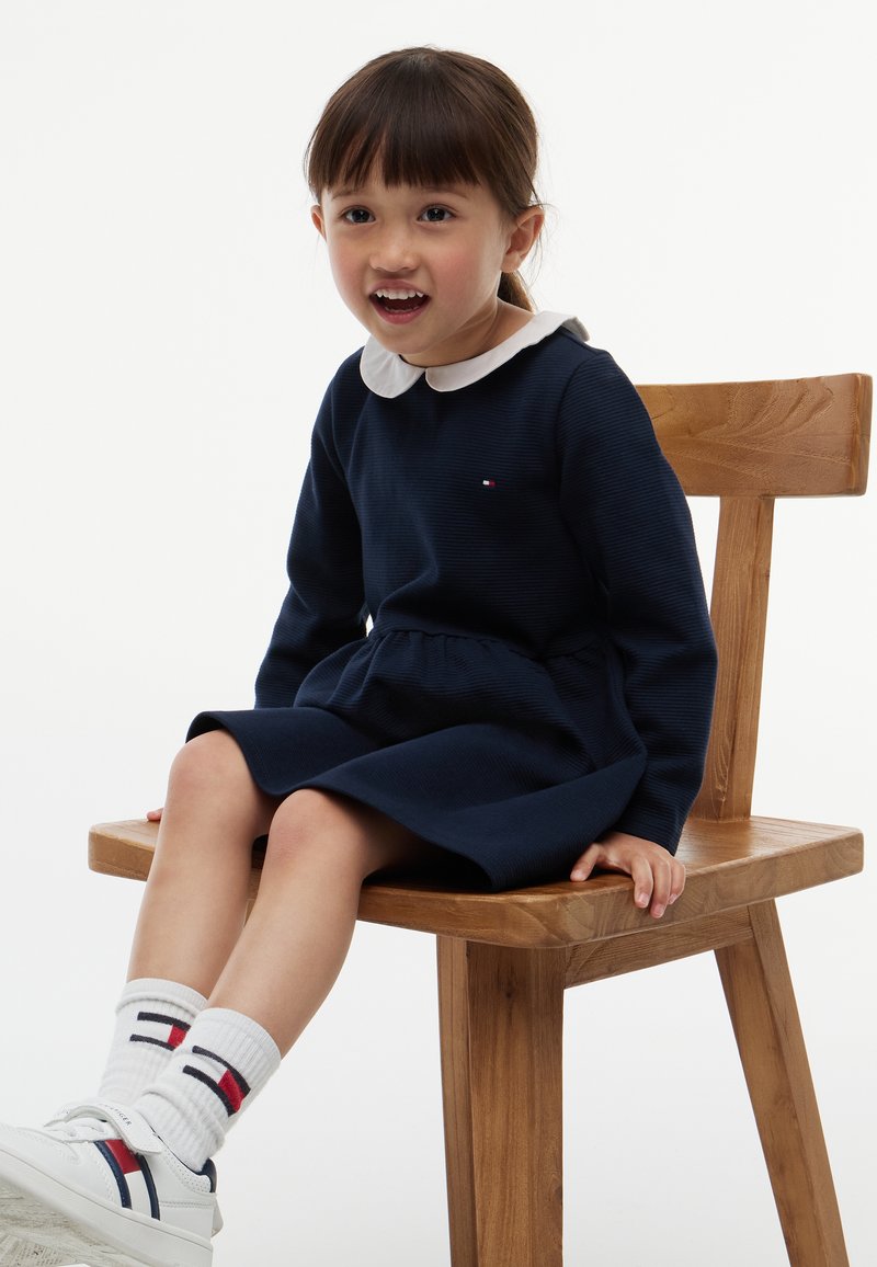 Jeune fille souriante, portant une robe bleu marine avec un col blanc, des chaussettes blanches et des baskets, assise sur une chaise en bois devant un fond uni.