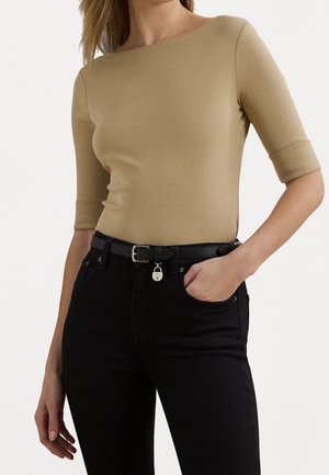 Vrouw draagt een beige nauwsluitend shirt met mouwen tot de ellebogen en zwarte broek met hoge taille, voorzien van een zwarte riem met een klein hangslot als charme.