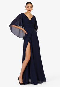 Frau, die ein marineblaues Abendkleid mit transparenten Kapuzenärmeln und einem hohen Schlitz trägt, kombiniert mit schwarzen High Heels.