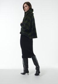 Groene en zwarte geruite oversized jas met hoge kraag, gecombineerd met een aansluitende zwarte rok en kniehoge zwarte laarzen met hak.