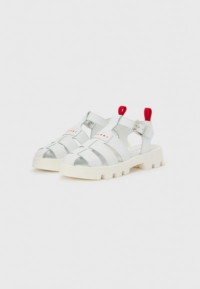 Λευκά σανδάλια με χοντρή σόλα, πολλαπλές λουρίδες, ασημένια αγκράφα, κόκκινες θηλιές στη φτέρνα και διακριτικό κόκκινο λογότυπο σε απλό ανοιχτό φόντο.