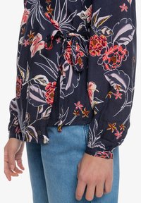 Blusa floreale in tessuto blu scuro, con vivaci fiori rossi e rosa. Legata in vita, con maniche lunghe e polsini con bottoni. Jeans azzurri.