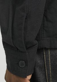 Poignet de veste tissé noir avec fermeture à bouton, présentant des détails de couture visibles et un tissu texturé ; à côté se trouve un denim foncé avec des coutures jaunes.