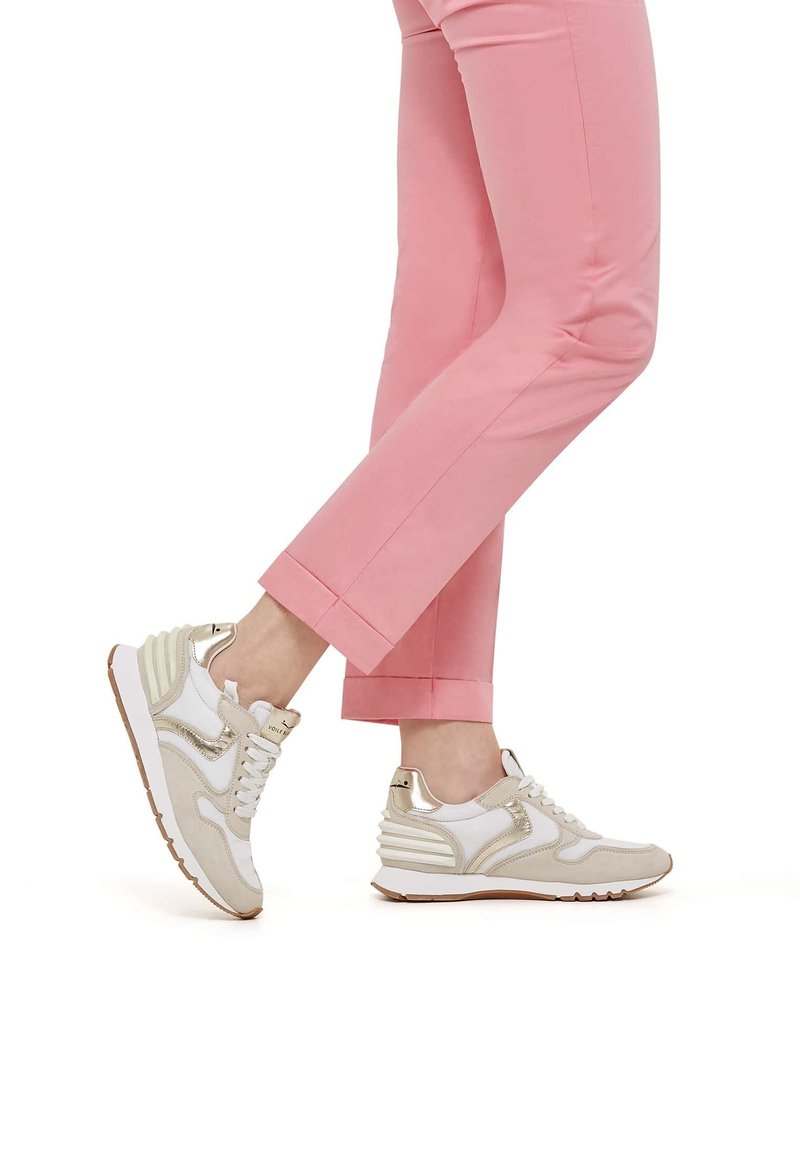 Leichte Sneakers in Weiß und Beige mit metallischen Akzenten. Das Design verfügt über eine gepolsterte Sohle und geschichtete Seitenpaneele. Getragen mit rosa Hosen.