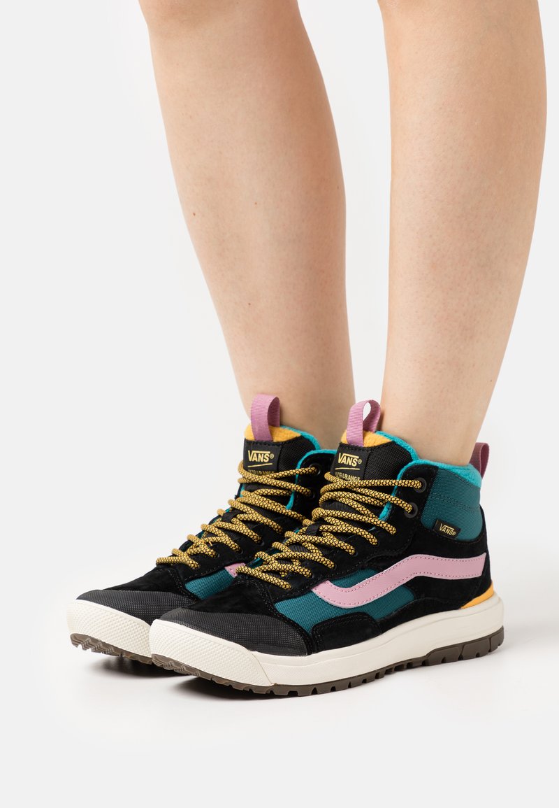 Zapatillas altas con un diseño negro y verde azulado, con acentos rosas y materiales texturizados. Cadenas doradas y suela de goma.