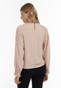 Lys beige bluse med smocked-detaljer på skuldre og ærmer, nøglehul-lukning bagpå og blødt draperet stof. Optrukne ærmer.