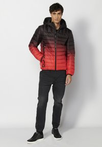 Giacca imbottita con una sfumatura dal nero al rosso, caratterizzata da una chiusura con zip, cappuccio e stampa del logo lungo il lato. Indossata con jeans scuri e sneakers.