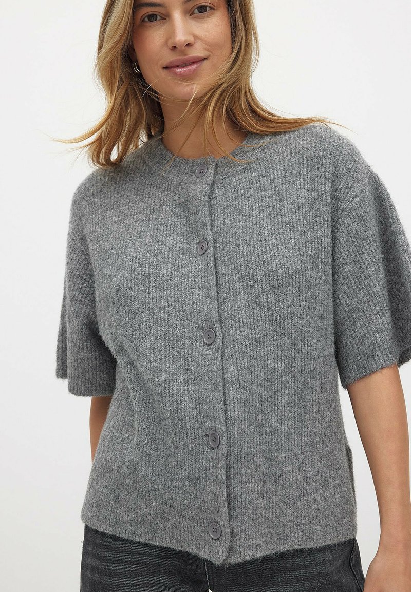 Pilkas trumpas rankovėmis kardiganas, pagamintas iš tekstūruotos siūlės, su penkiomis sagomis priekyje ir suapvalintu kaklu.