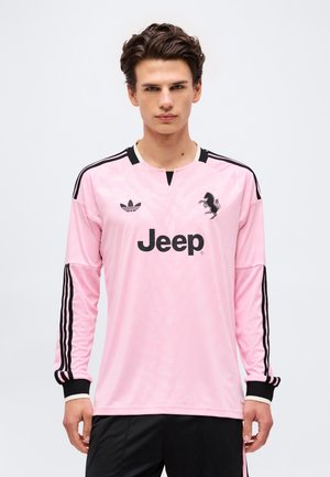 Młody mężczyzna w różowej długorękawowej sportowej koszulce Adidas z czarnym logo Jeep i czarnymi paskami na rękawach.