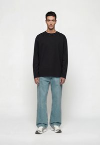 Άνδρας που στέκεται, φοράει μαύρο μπλουζάκι με μακριά μανίκια, ανοιχτό μπλε τζιν με φαρδιά μπατζάκια και λευκά αθλητικά παπούτσια, μπροστά σε απλό ανοιχτό γκρι φόντο.