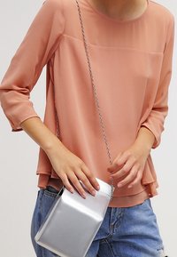 Bluzka w kolorze brzoskwiniowym, lekka, z rękawami 3/4 i warstwowym dołem. Srebrna torebka na ramię z gładką, elegancką powierzchnią i łańcuszkowym paskiem.