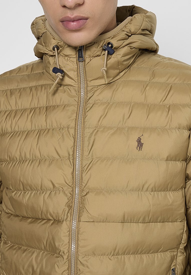 Mann trägt eine sandfarbene Steppjacke mit Kapuze und zentralem Reißverschluss, die auf der Brust ein kleines dunkles Polo-Spieler-Logo zeigt.