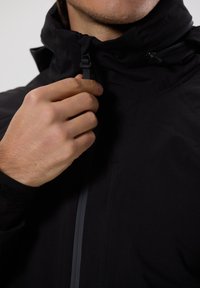 Černá bunda s vysokým límcem, se zipem a rukou držící zip. Hladká tkanina s matným povrchem.