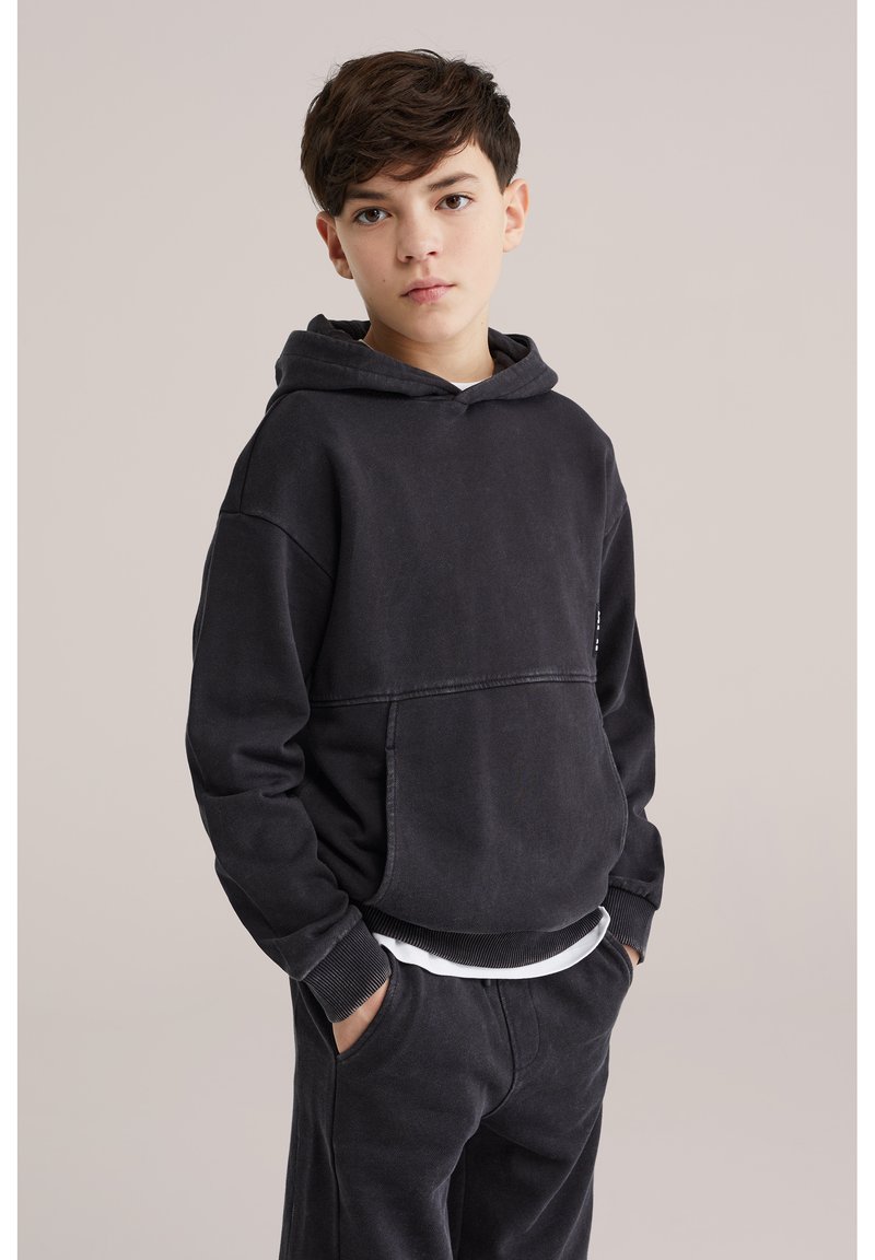 Jongen draagt een zwarte hoodie en bijpassende broek met handen in de zakken, staand tegen een effen lichte achtergrond.