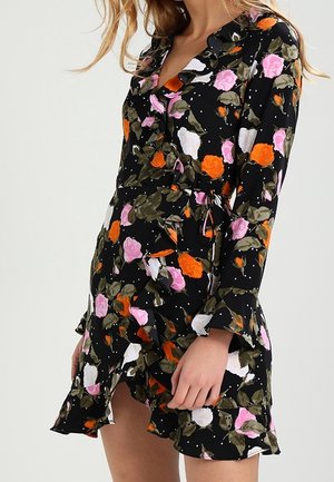Woman wearing a black wrap dress with orange, pink, and white floral print, long sleeves, and ruffled hem and cuffs.