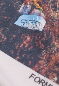 T-shirt en coton présentant un graphisme multicolore avec des tons terreux bruns, des accents bleus, et un texte manuscrit près du bas.