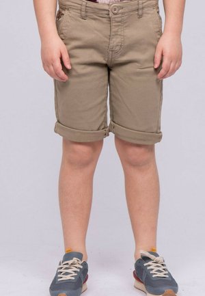 Enfant portant un short beige retroussé et des baskets bleues, debout sur un fond uni.