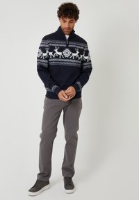 Marineblauer gestrickter Pullover mit einem Viertelreißverschluss, verziert mit weißen Rentiermustern und Schneeflocken. Graue Hosen und dunkle Schuhe runden das Outfit ab.