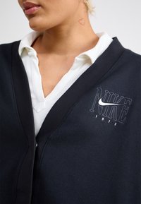 Närbild på person i vit skjorta med krage och svart Nike-kofta med logotyp och texten "1972", hjärttatuering synlig på nacken.