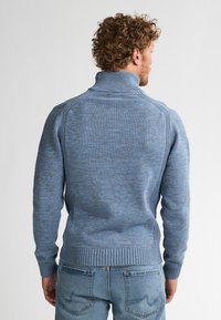 Hombre con cabello rizado, vistiendo un suéter azul de cuello alto de punto y jeans azul claro, de pie de espaldas frente a un fondo liso.