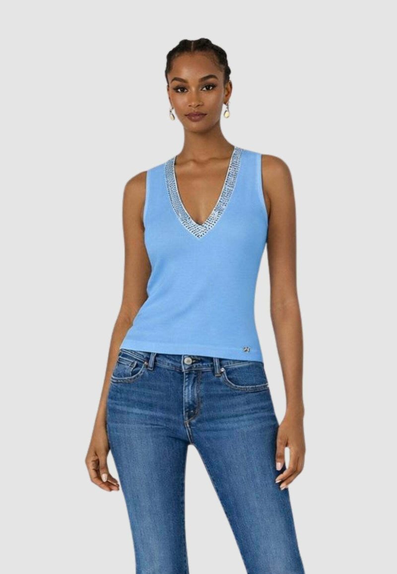 Donna che indossa un top senza maniche color azzurro con scollatura a V e dettagli argentati abbinato a jeans di denim blu, su uno sfondo semplice.