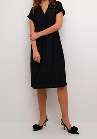 Vestido negro hasta la rodilla con mangas cortas, que presenta detalles de ojal y un corte relajado. Combinado con tacones descalzos negros adornados con una flor.